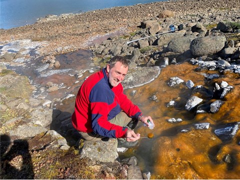 Michael Kühl indsamler biofilm i en 55 grader varm kilde i Knighton Bugt, Blosseville Kyst, Østgrønland. I kilden dannes flere cm tykke orange belægninger af biofilm, der opbygges af cyanobakterier indlejret i slimstoffer, som de udskiller. (Foto: Christiane Leister; fra Kühl og Colgan, 2025)
